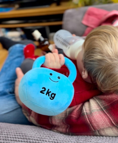 Baby on sofa drinking milk while holding Hazza Toyz Mystic Blue kettlebell, enjoying a cosy and playful moment