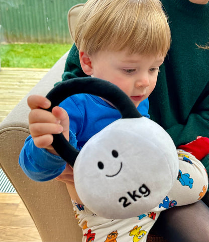 Baby sitting on mum's lap on the sofa, holding Hazza Toyz Lamby Black and White kettlebell, enjoying a cosy moment and playful interaction