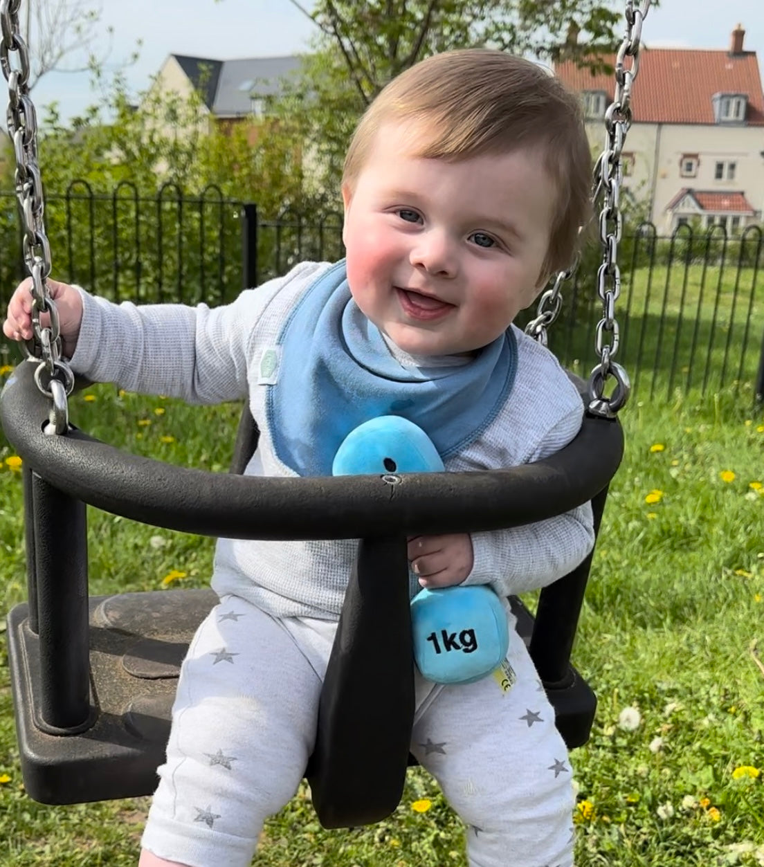 Baby in park swing holding Hazza Toyz Mystic Blue dumbbell, enjoying outdoor sensory play and motor skill development