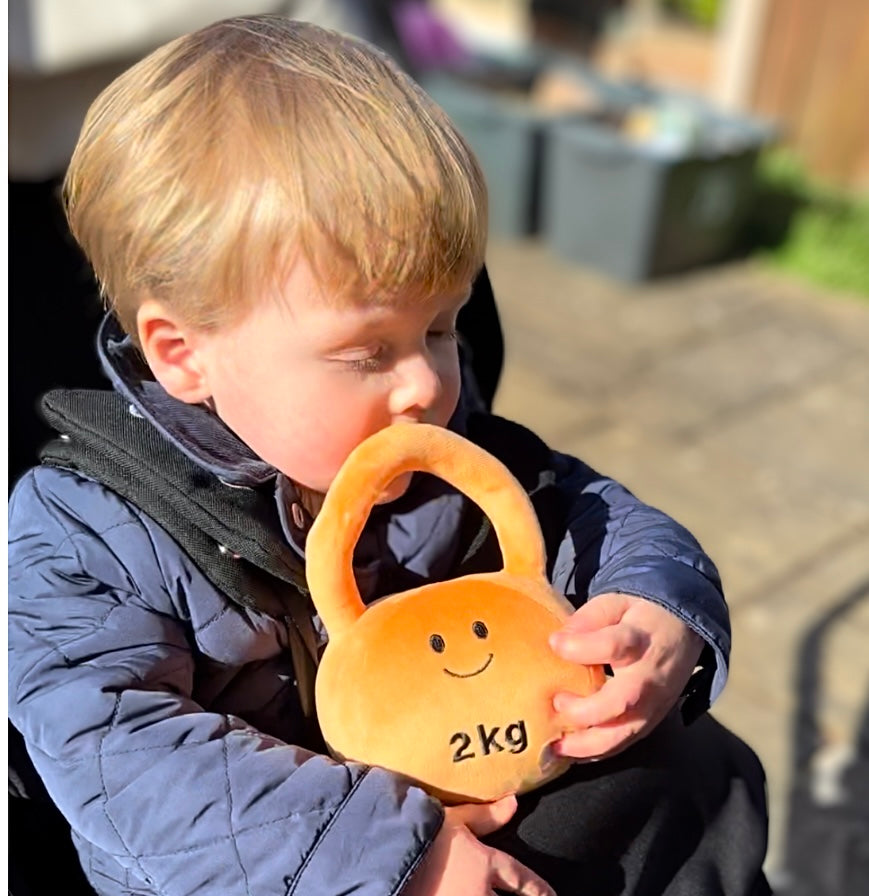 Baby in pram hugging Hazza Toyz Apricot Orange kettlebell, enjoying sensory comfort and play during an outdoor stroll