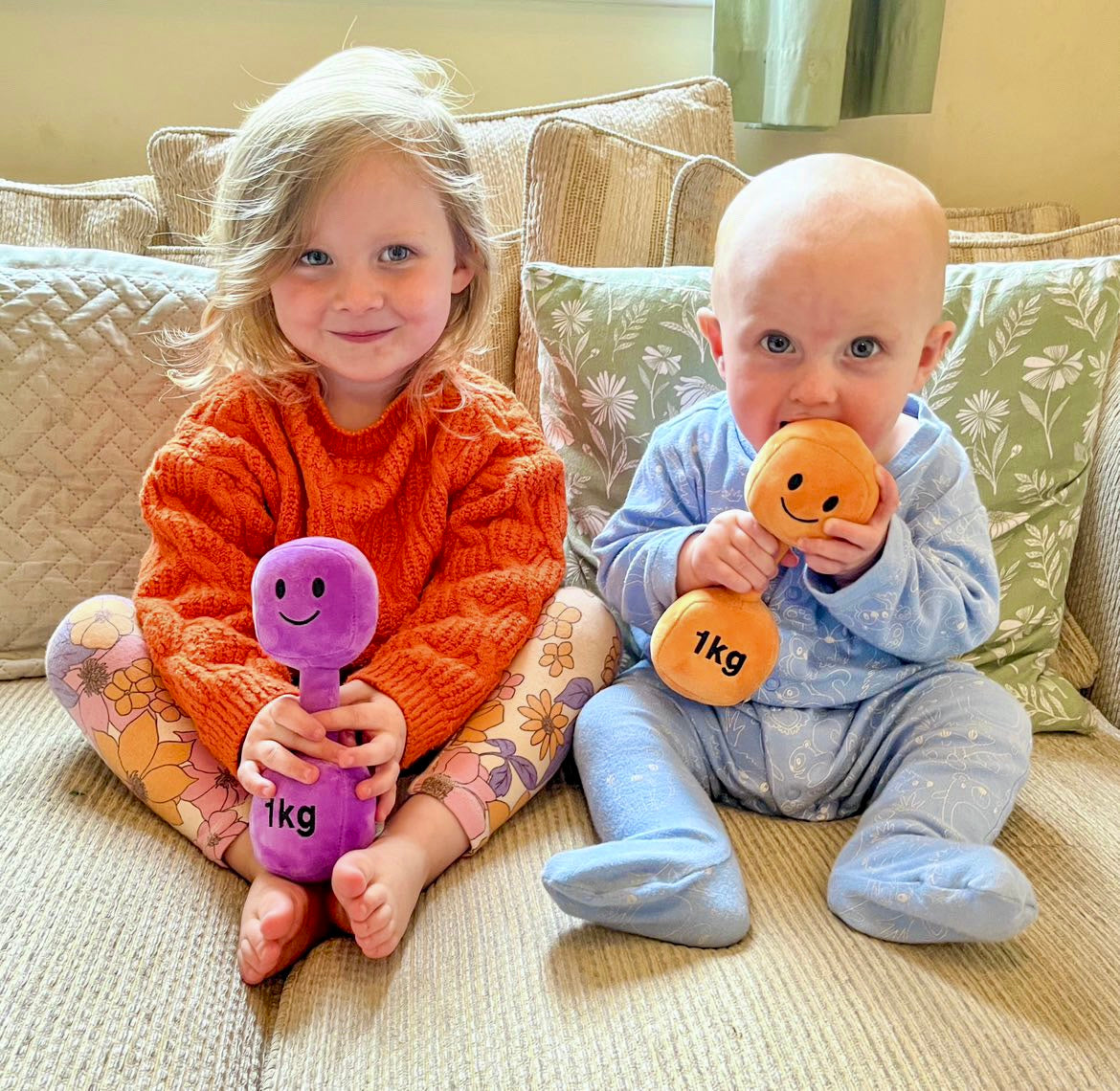 Baby sister and brother each holding a Hazza Toyz dumbbell—one in Apricot Orange and one in Lavender Purple—sharing playful sensory development and motor skill activities