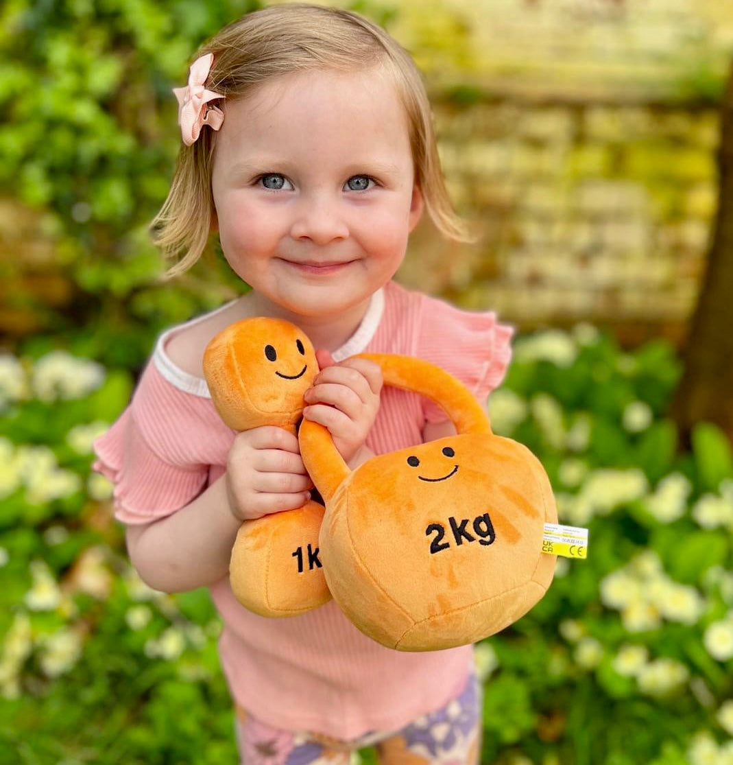 Baby smiling at camera in garden, holding Hazza Toyz Apricot Orange dumbbell and kettlebell, enjoying outdoor sensory play