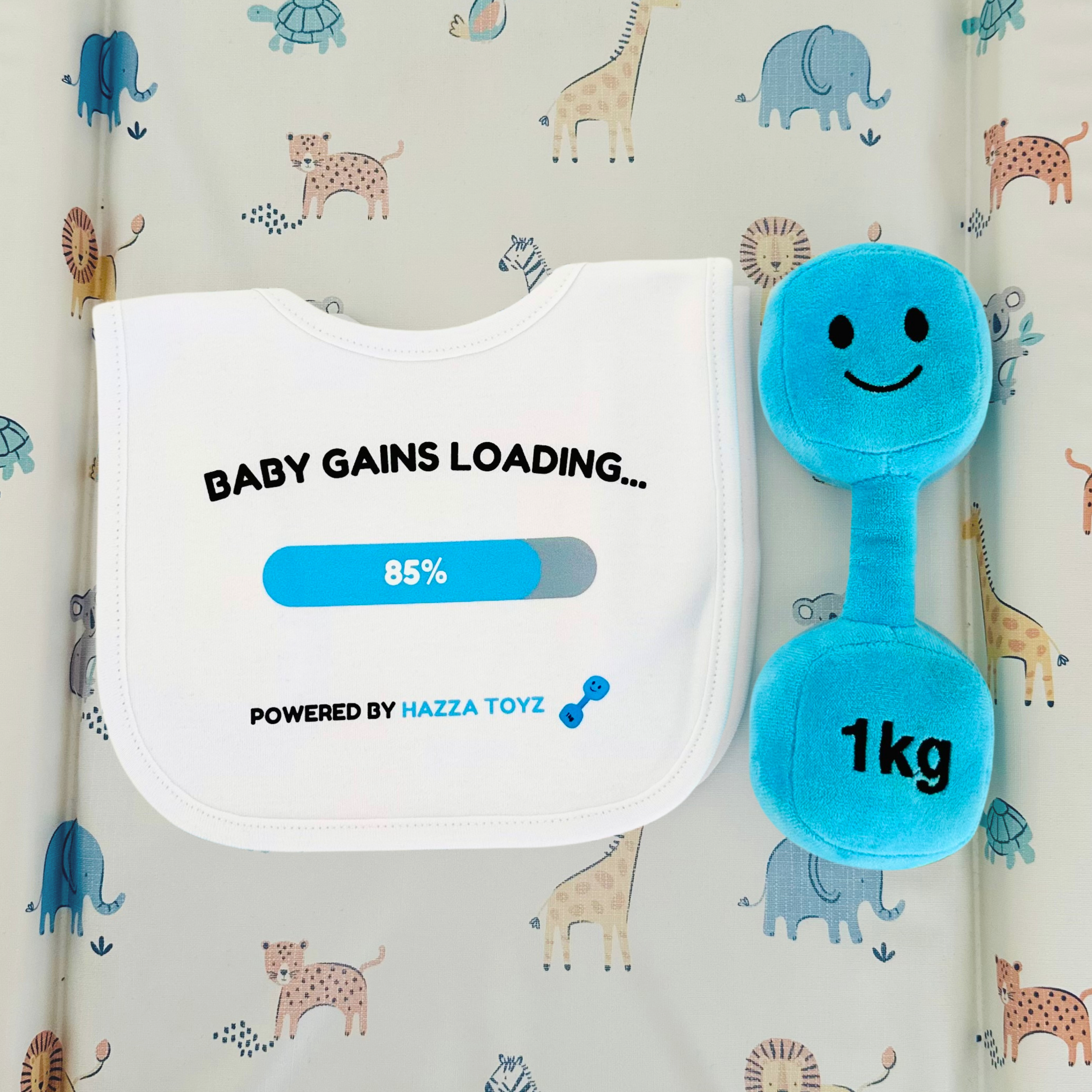 White “Baby Gains Loading…” bib at 85% with a blue plush 1kg dumbbell toy, displayed on a baby animal print mat.