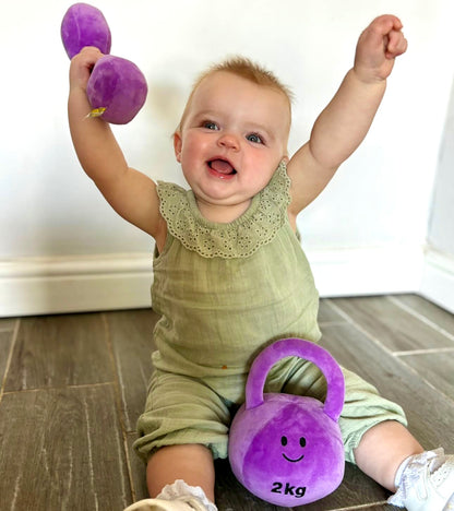 Baby happily lifting Hazza Toyz Lavender Purple dumbbell with kettlebell nearby, engaging in sensory play and motor skill development