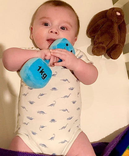 Little baby in cot holding Hazza Toyz Mystic Blue dumbbell, with brown plush bunny in background, enjoying sensory play and comfort