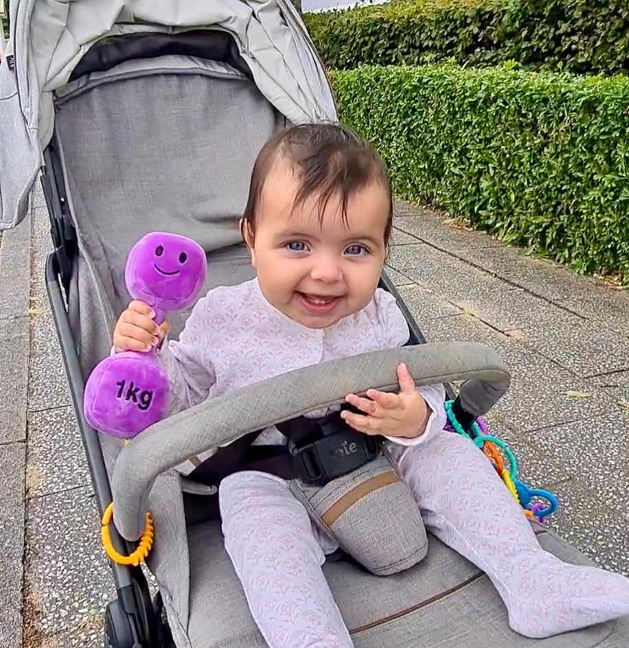 Happy baby in pram holding Hazza Toyz Lavender Purple dumbbell, enjoying sensory play while out on a stroll