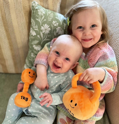 Smiling siblings cuddling with Hazza Toyz Apricot Orange dumbbell and kettlebell, holding baby toys and enjoying playful bonding time