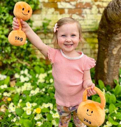 Toddler in garden playing with Hazza Toyz Apricot Orange plush dumbbell and kettlebell, enjoying outdoor sensory play and motor skill development