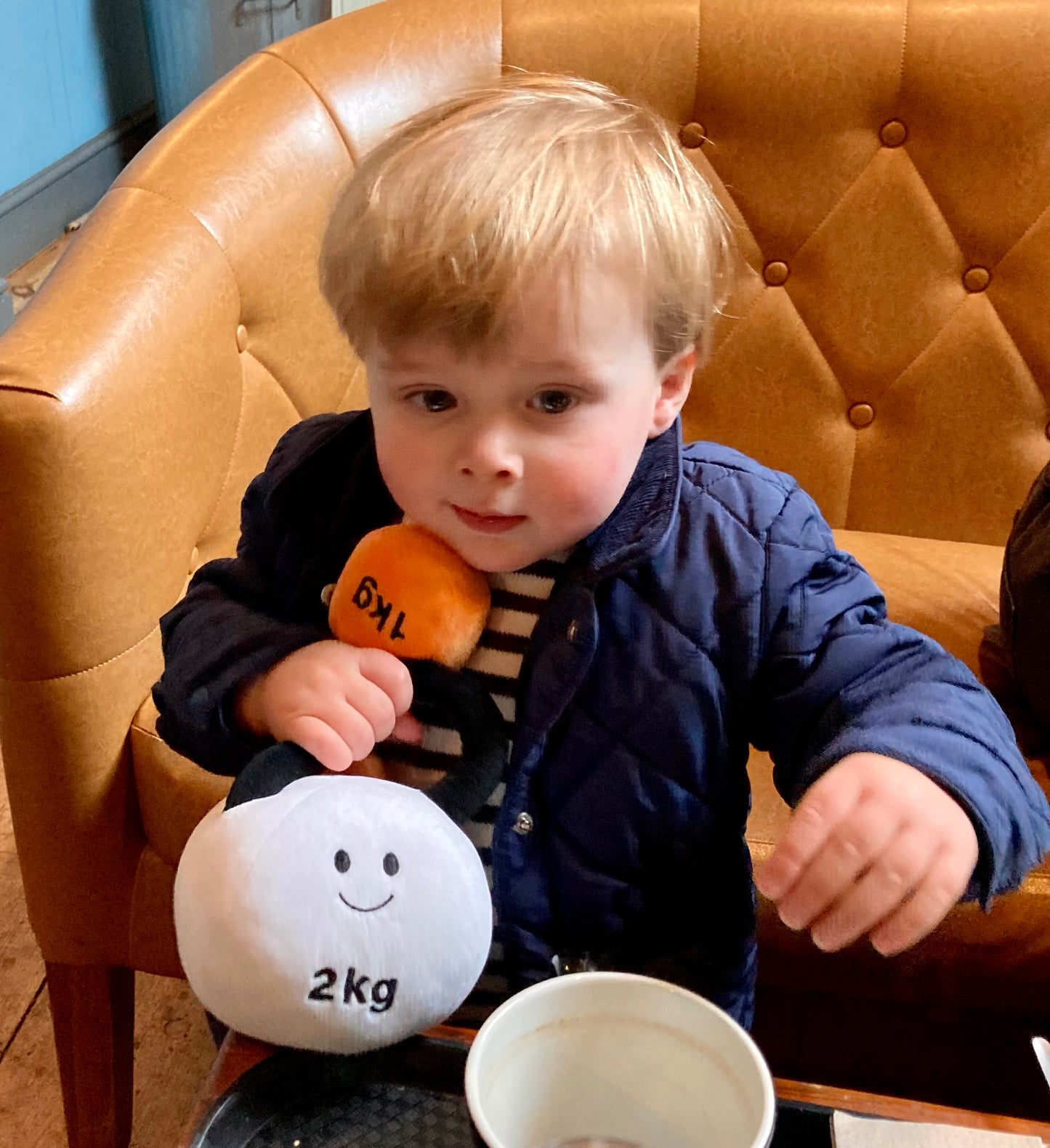 Toddler holding Hazza Toyz Lamby Black and White kettlebell and Apricot Orange dumbbell in coffee shop, enjoying playful interaction in a cosy setting