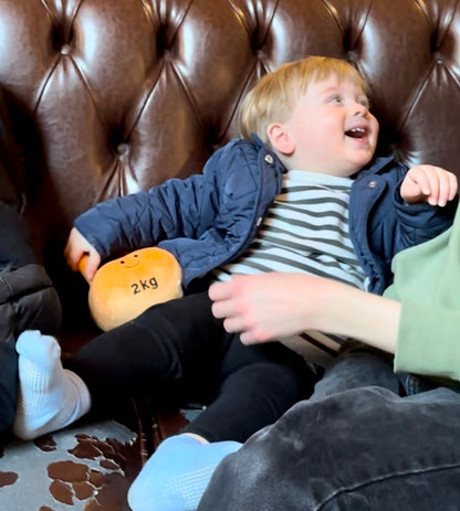 Toddler on leather sofa in coffee shop smiling at mum holding Hazza Toyz Apricot Orange kettlebell, enjoying playful interaction in a cosy setting