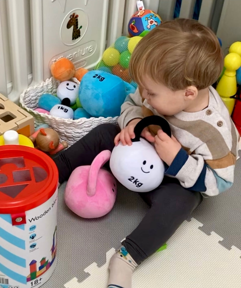 Toddler sitting on playmat next to Hazza Toyz items, playing with Lamby Black and White kettlebell, enjoying sensory play and developing motor skills