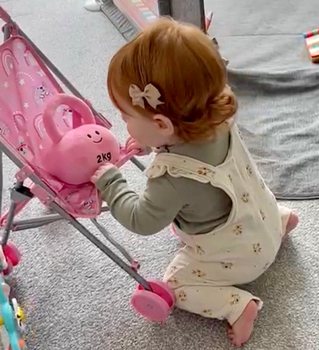 Toddler playing with Hazza Toyz Candy Chateau Pink kettlebell in pram, enjoying sensory play and motor skill development on the go