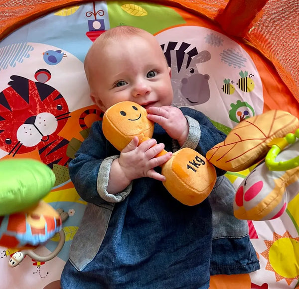 Baby lying on colourful play mat chewing Apricot Orange plush 1kg dumbbell toy
