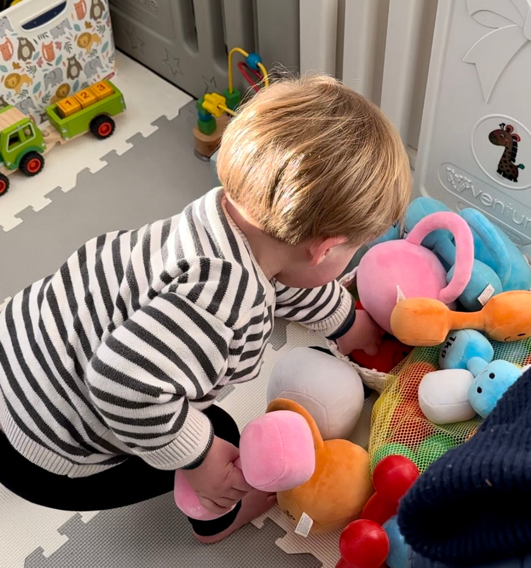 Toddler leaning over a pile of baby toys to grab a Hazza Toyz item, showing curiosity and active engagement in playtime