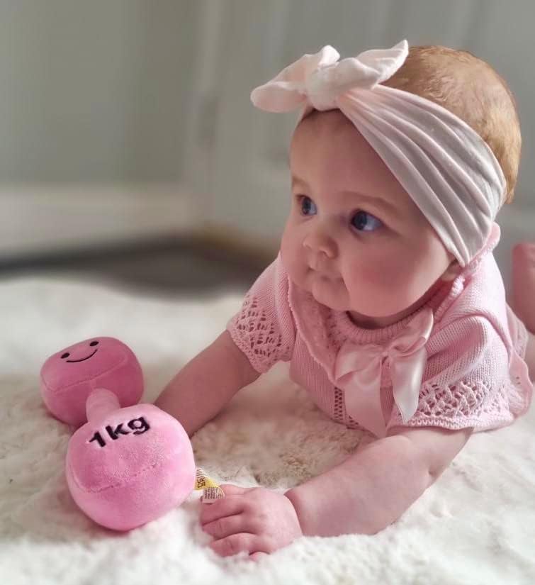 Baby girl wearing a white headband, reaching out for a Candy Chateau Pink dumbbell, engaging in curious play with Hazza Toyz fitness-inspired toys for toddlers.