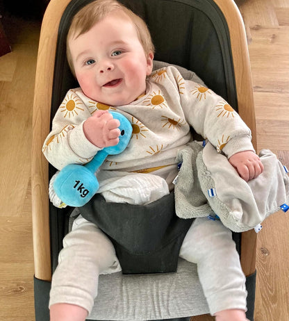 Happy baby holding a Mystic Blue dumbbell while sitting in a car seat, enjoying travel-friendly fun with Hazza Toyz fitness-inspired toys for toddlers.