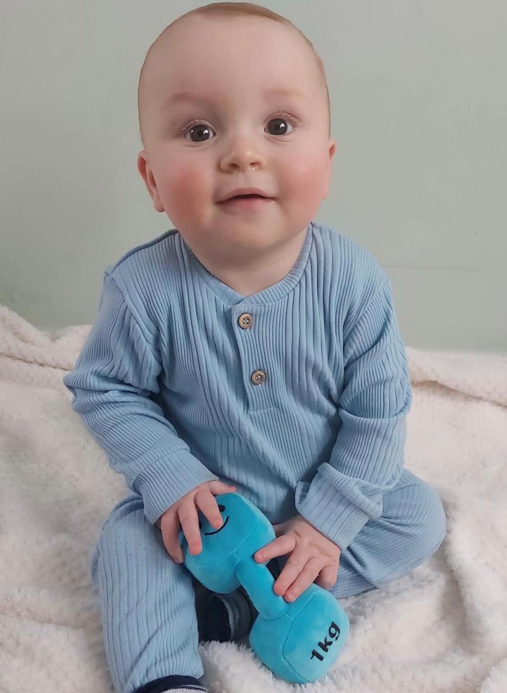 Baby sitting up with a cute smile, holding a Mystic Blue dumbbell, enjoying playful moments with Hazza Toyz fitness-inspired toys for toddlers.