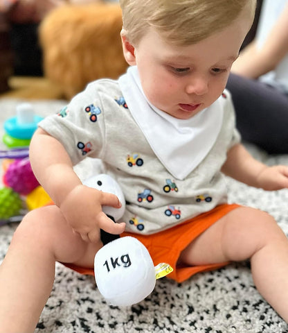 Baby wearing a white bib and holding a Lamby Black and White dumbbell, enjoying playtime with Hazza Toyz fitness-inspired toys for toddlers.