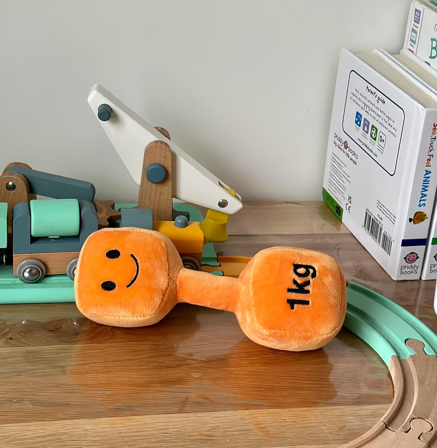 Hazza Toyz Apricot Orange dumbbell placed next to a toy train set, combining soft sensory play with classic wooden toys for toddlers